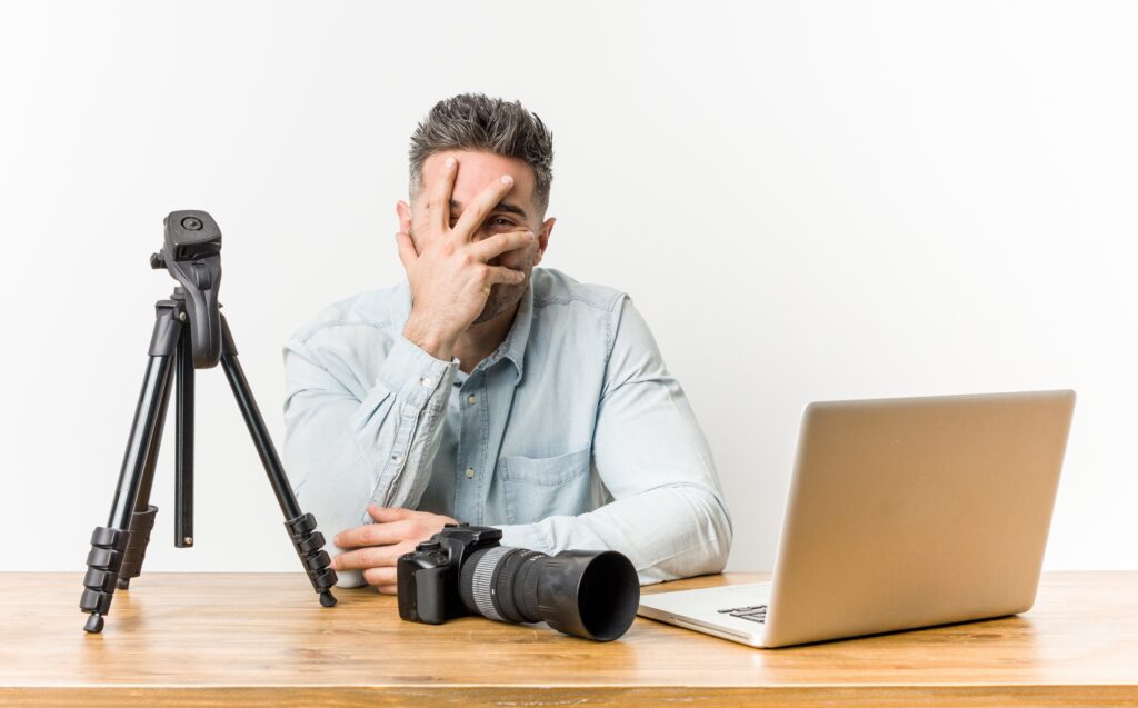 Muž sedí za dreveným stolom a má pred sebou notebook, profesionálnu kameru a tripod. Chytá sa za hlavu a zakrýva si tvár.